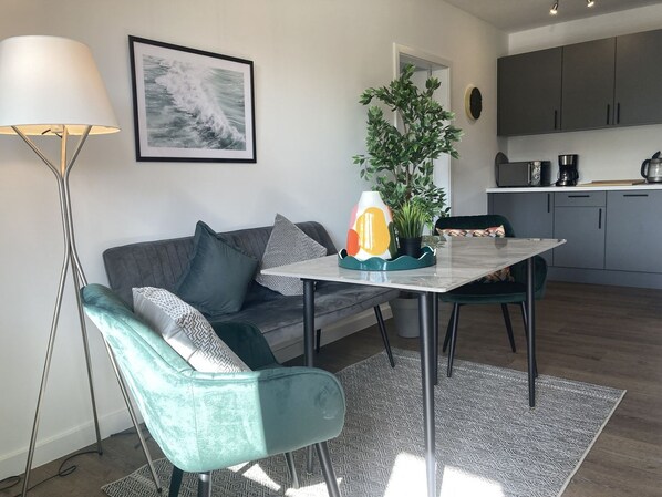 A modern living space with a sofa, a table, a floor lamp, a potted plant, and a kitchen area in the background.