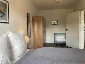 A bedroom with a bed, a wooden wardrobe, a bedside table with a lamp, and a bunk bed in the background.