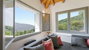 Una sala de estar moderna con un sofá gris, cojines rojos y grises, y ventanas amplias que ofrecen vistas a un paisaje verde y montañas.