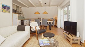 Una sala moderna con un sofá, sillas, una mesa de centro y un televisor. Se observa un área de comedor con una mesa y sillas al fondo.