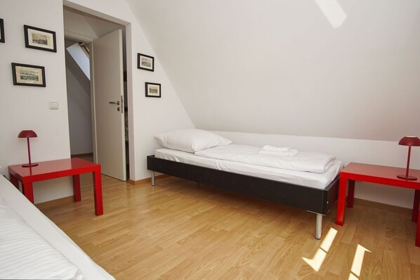 Un dormitorio con cama alta, dos mesitas de noche rojas, una puerta blanca y cuadros en la pared.