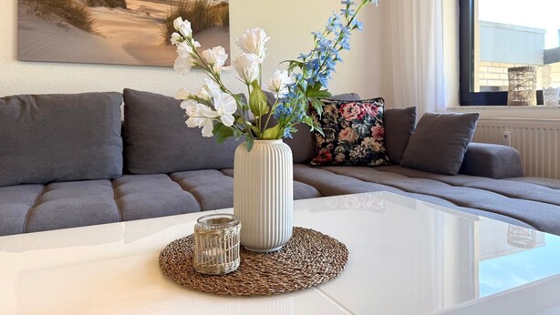A living room with a white coffee table, a vase with white and blue flowers, a woven coaster, and a painting of a beach scene.