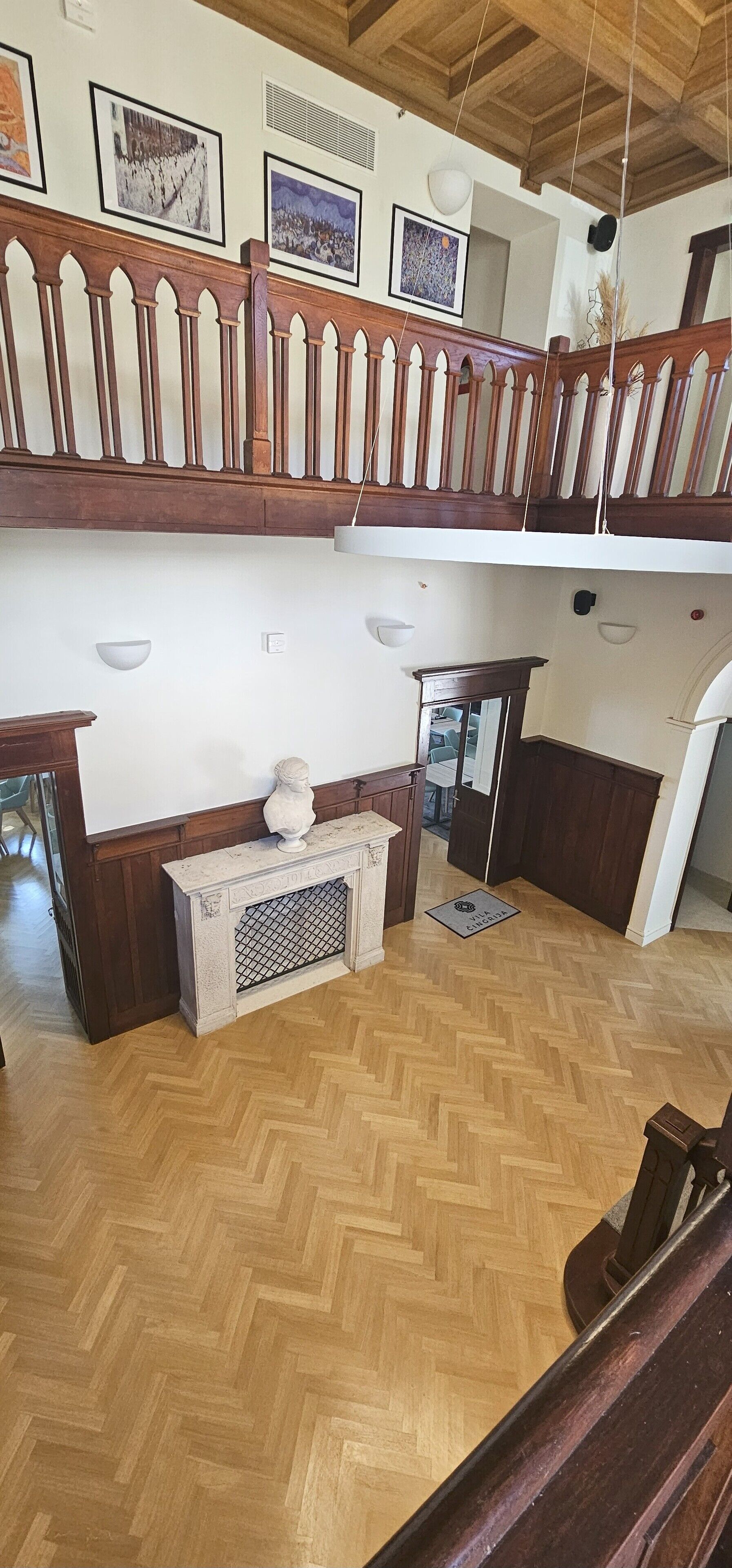 A spacious room with a wooden herringbone floor, a stone fireplace, and a balcony with a wooden railing.