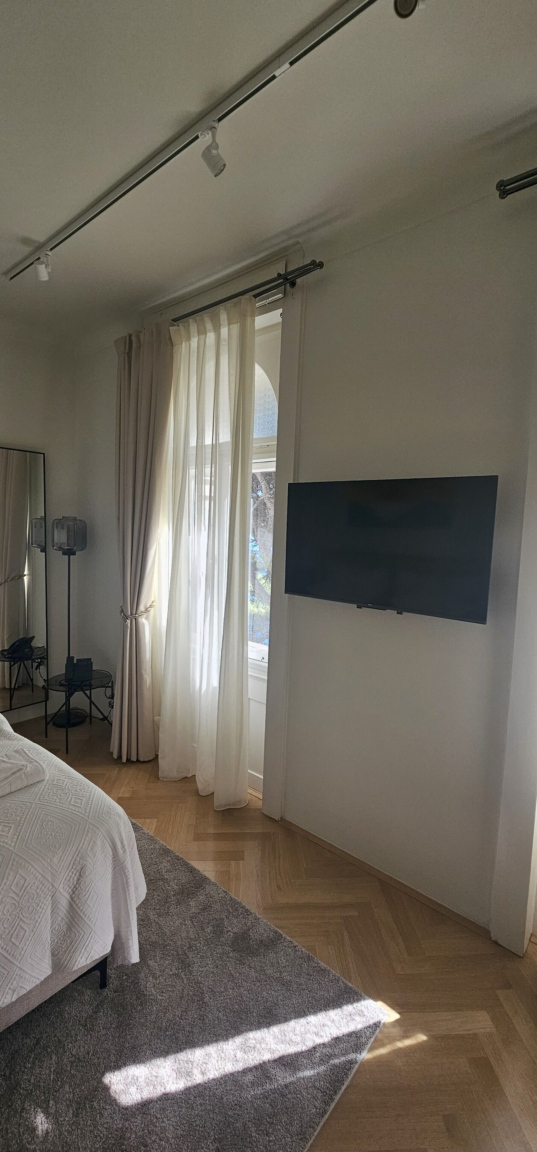 Un dormitori amb un llit, un televisor fixat a la paret, escriptori amb cadira i finestra amb cortines.