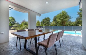 An outdoor dining area with a pool view, featuring a wooden table and chairs, a bowl of fruit, and a glass pitcher. - Stunning home in Dekani with private swimming pool, can be inside or outside (Dekani)