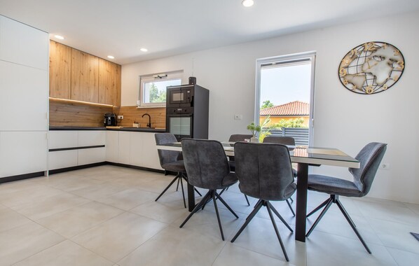 A modern kitchen with white cabinets, a wooden accent wall, and a dining area with four grey chairs and a glass table.