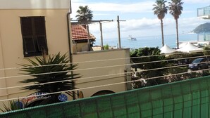 A balcony with a round table and two chairs, overlooking a coastal area with palm trees and a building with a tiled roof. - Raffaello Beach Apartment Savona, 50 m from the sea, WIFI, AC, equipped kitchen (Savona)