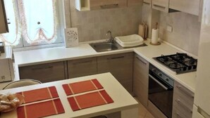 A modern kitchen with a built-in gas stove, a sink, and a microwave. There are two bar stools and a window with curtains. - Raffaello Beach Apartment Savona, 50 m from the sea, WIFI, AC, equipped kitchen (Savona)