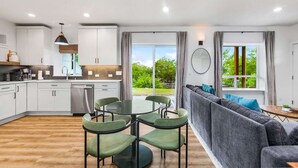 A modern kitchen with white cabinets, a dining area with a round table and green chairs, and a gray sofa with blue cushions.