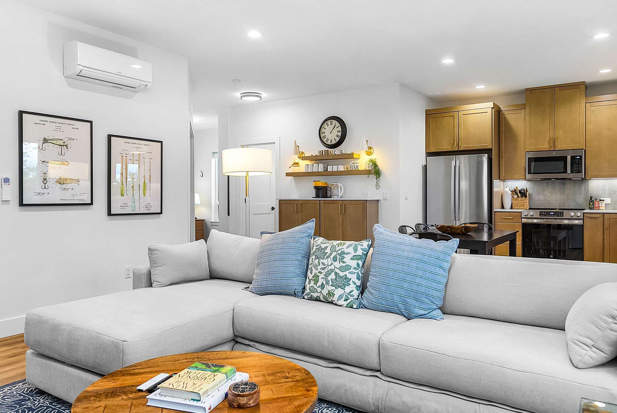 A modern living room with a grey sofa, a coffee table, and a kitchen area with wooden cabinets and stainless steel appliances.