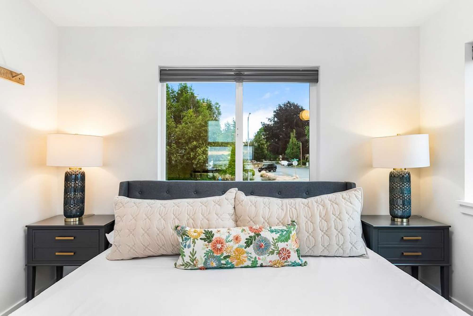 A bedroom with a large bed, two bedside tables with lamps, a window with a view of trees and a road, and a floral pillow on the bed.