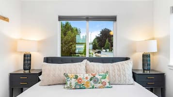A bedroom with a large bed, two bedside tables with lamps, a window with a view of trees and a road, and a floral pillow on the bed.