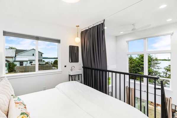 Une chambre avec un grand lit, une vue sur une maison et sur l’eau, une petite salle de bain et un balcon avec une balustrade.