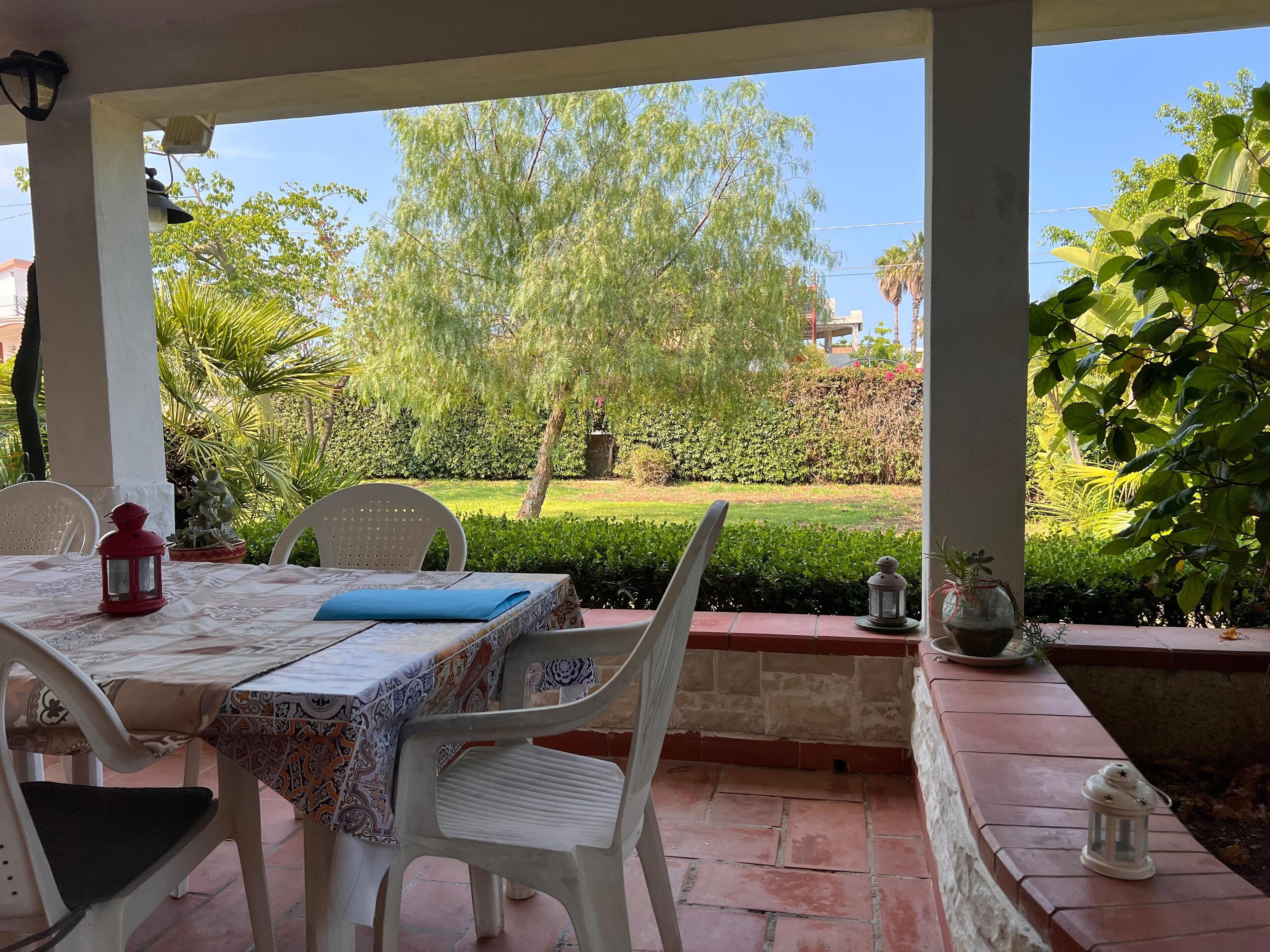 Eine überdachte Terrasse mit Tisch für zwei Personen, umgeben von Stühlen. Im Hintergrund ist ein kleiner Garten mit Hecke und Bäumen zu sehen.