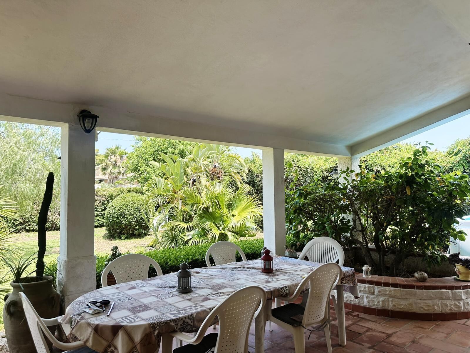 En overdækket terrasse med et spisebord, omgivet af stole, og udsigt til frodig vegetation.