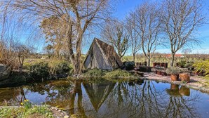 Ein rustikales Holzgebäude an einem Teich, umgeben von Vegetation, mit blauem Himmel.