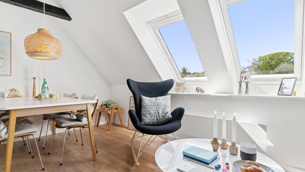 A modern loft bedroom with a sloped ceiling, large skylights, a round white table, and a dark armchair with a floral pillow.