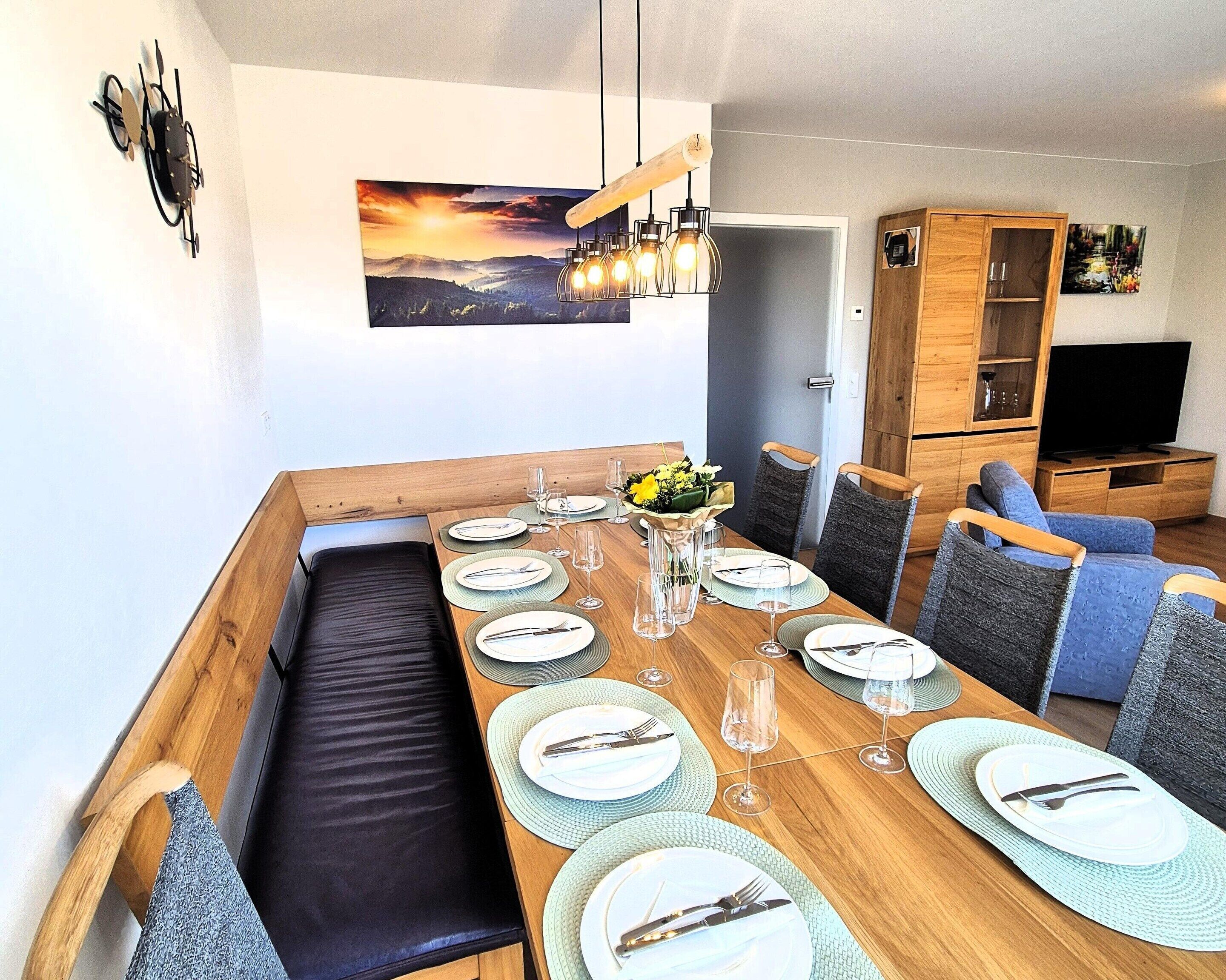 A dining area with a long wooden table set for six, surrounded by cushioned chairs, a bench, and a wooden cabinet with a television.
