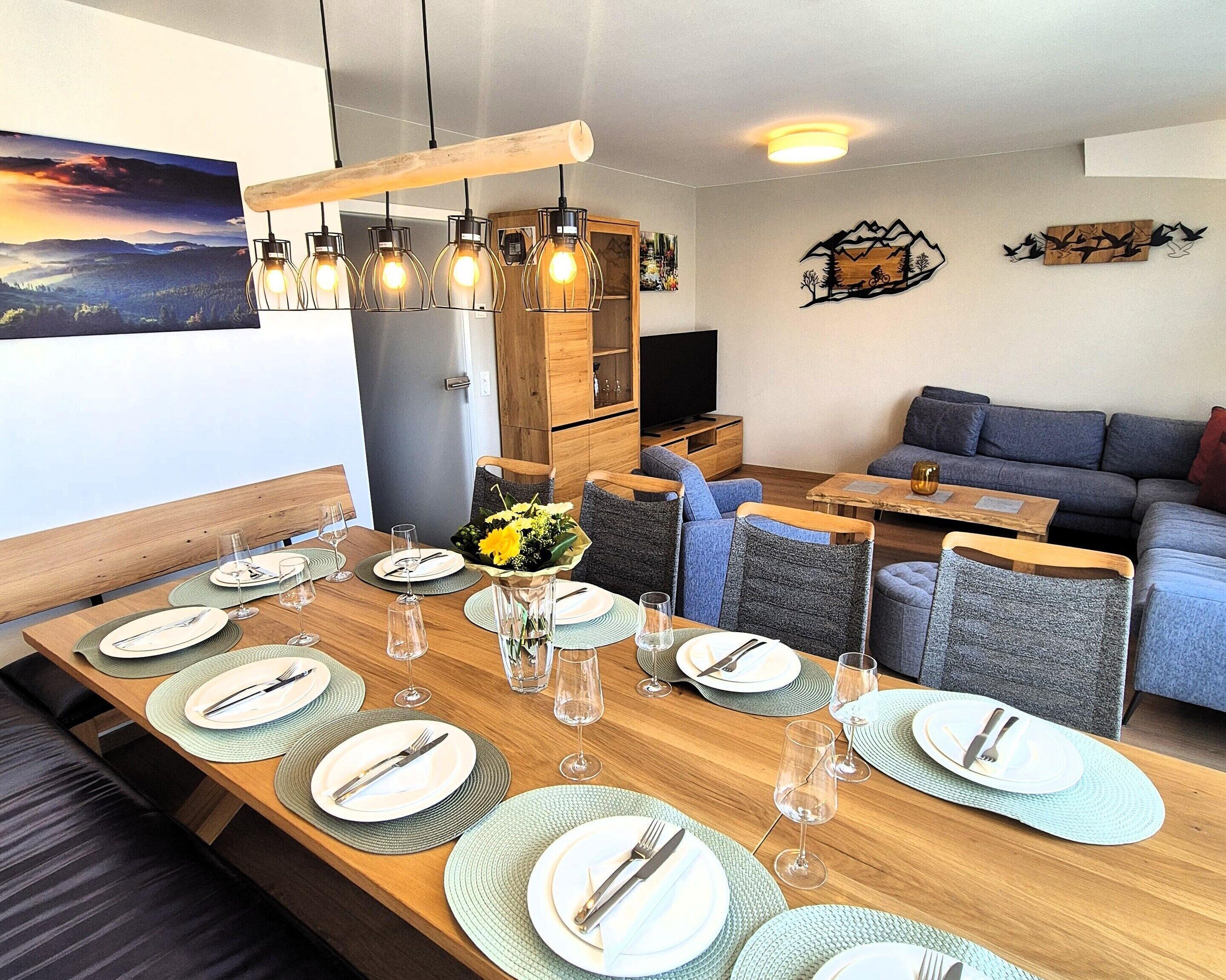 A dining area with a wooden table set for six, a sofa, and a TV in the background.