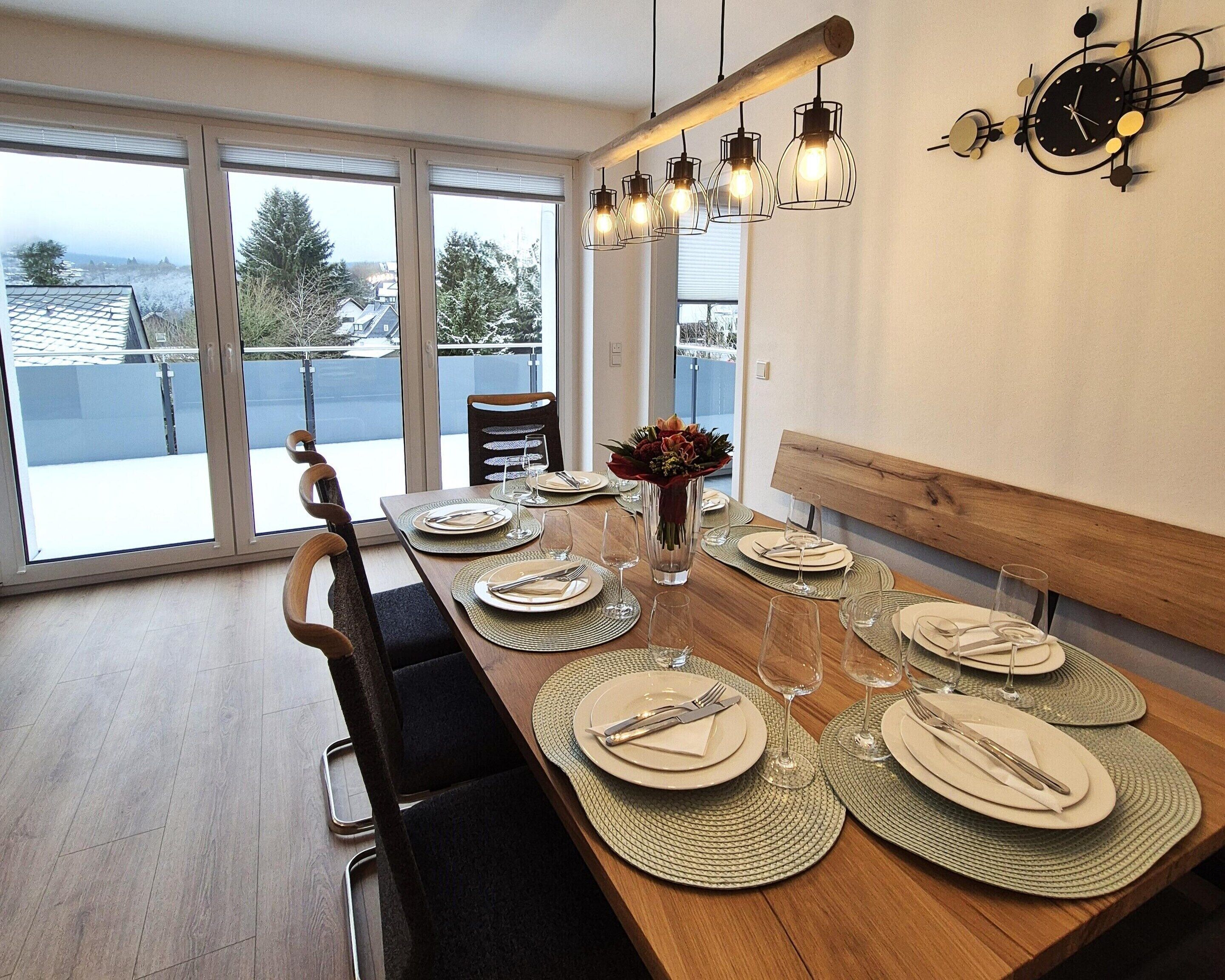 A dining area with a long wooden table set for six, large windows, and a view of the outside.