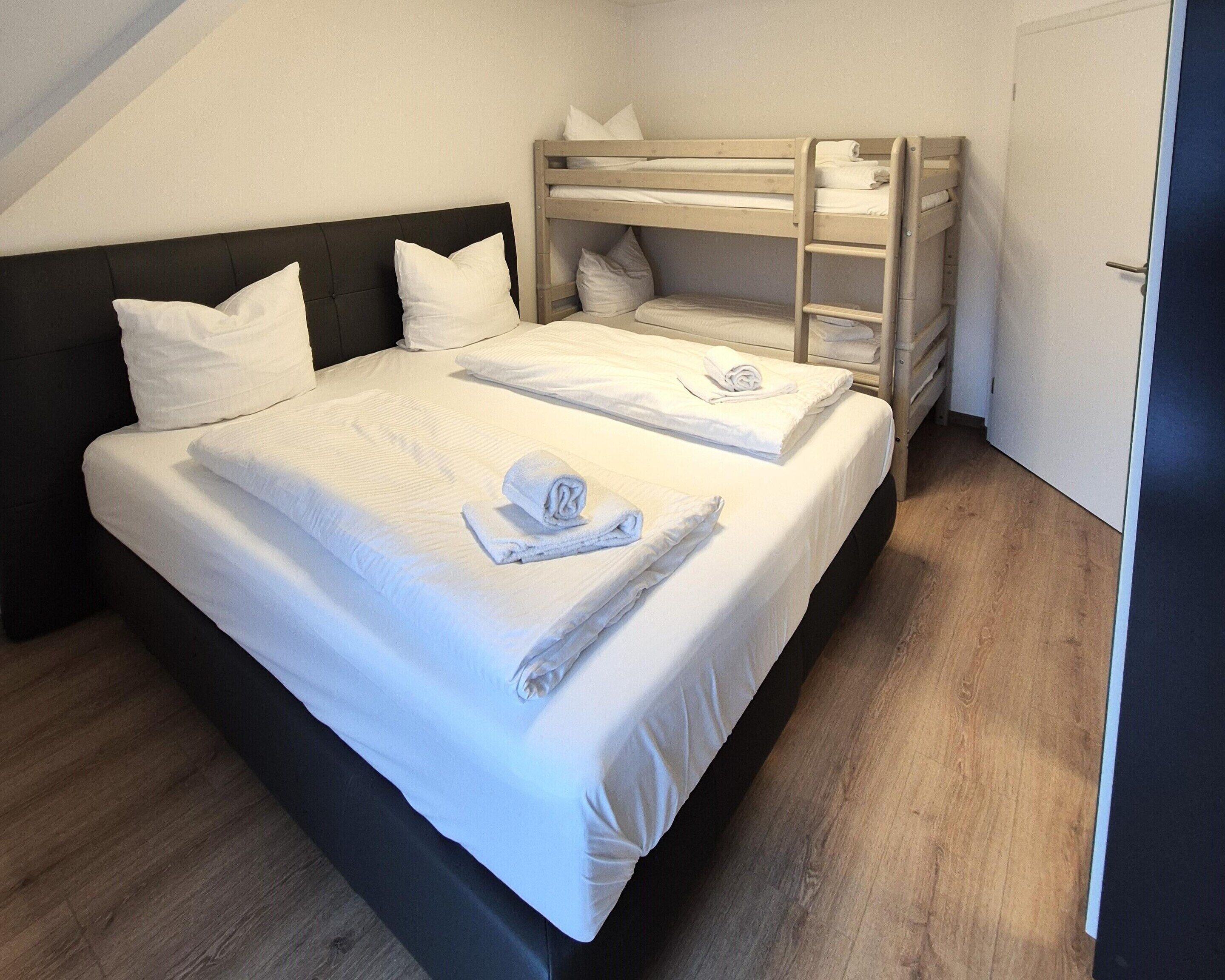 Una cama bien hecha con sábanas y toallas blancas, una litera con barandillas de madera y una puerta a la derecha.