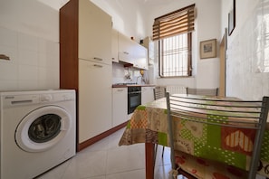 A compact kitchen with a Kenmore washer, a dining table with a patchwork tablecloth, and a window with blinds.
