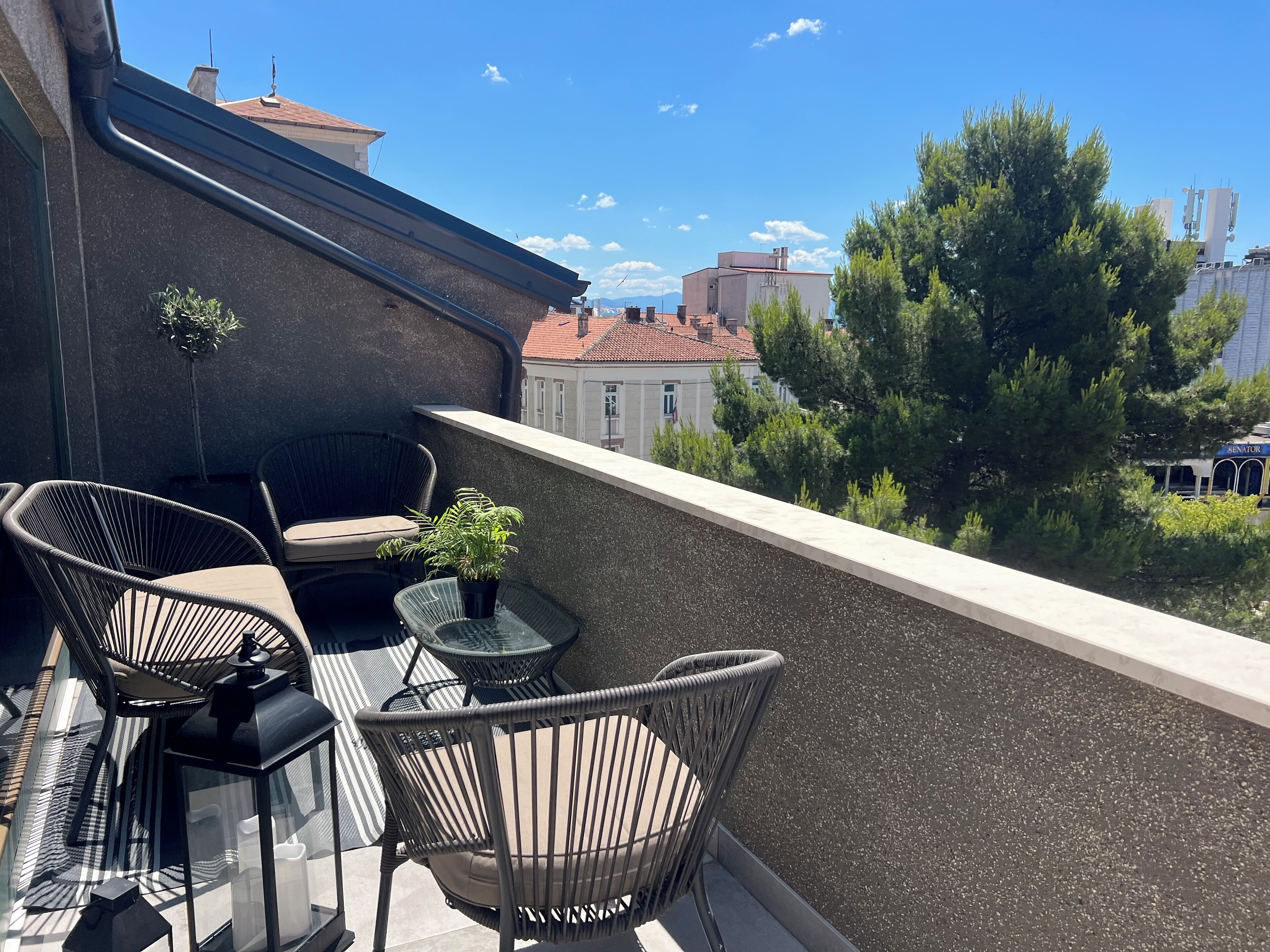A balcony with wicker chairs and a small table, a potted plant, and a view of buildings and trees.