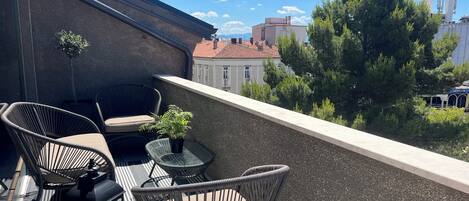 A balcony with wicker chairs and a small table, a potted plant, and a view of buildings and trees.