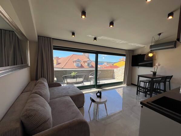 A modern living room with a sofa, a small table, and a balcony with a view of red-roofed buildings.