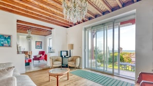 A spacious living room with a wooden ceiling, a chandelier, a large sliding glass door leading to a balcony, and a cozy seating area.