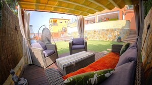 A covered outdoor seating area with wicker furniture, a stone wall, and a view of a yellow building. - Cozy house with pool (CT)
