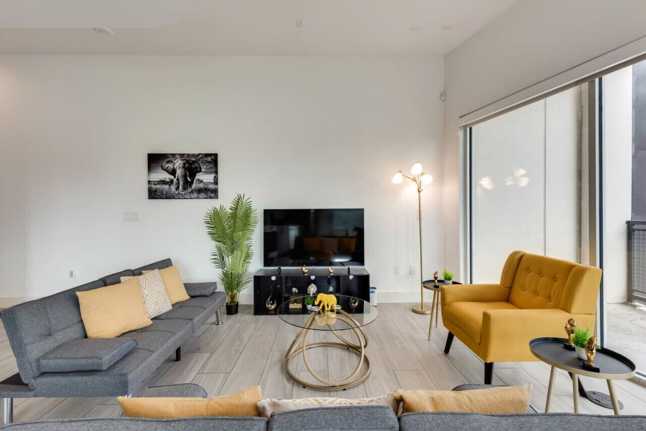 A modern living room with a grey sofa, a mustard-colored armchair, a flat-screen TV, a floor lamp, and a large window.