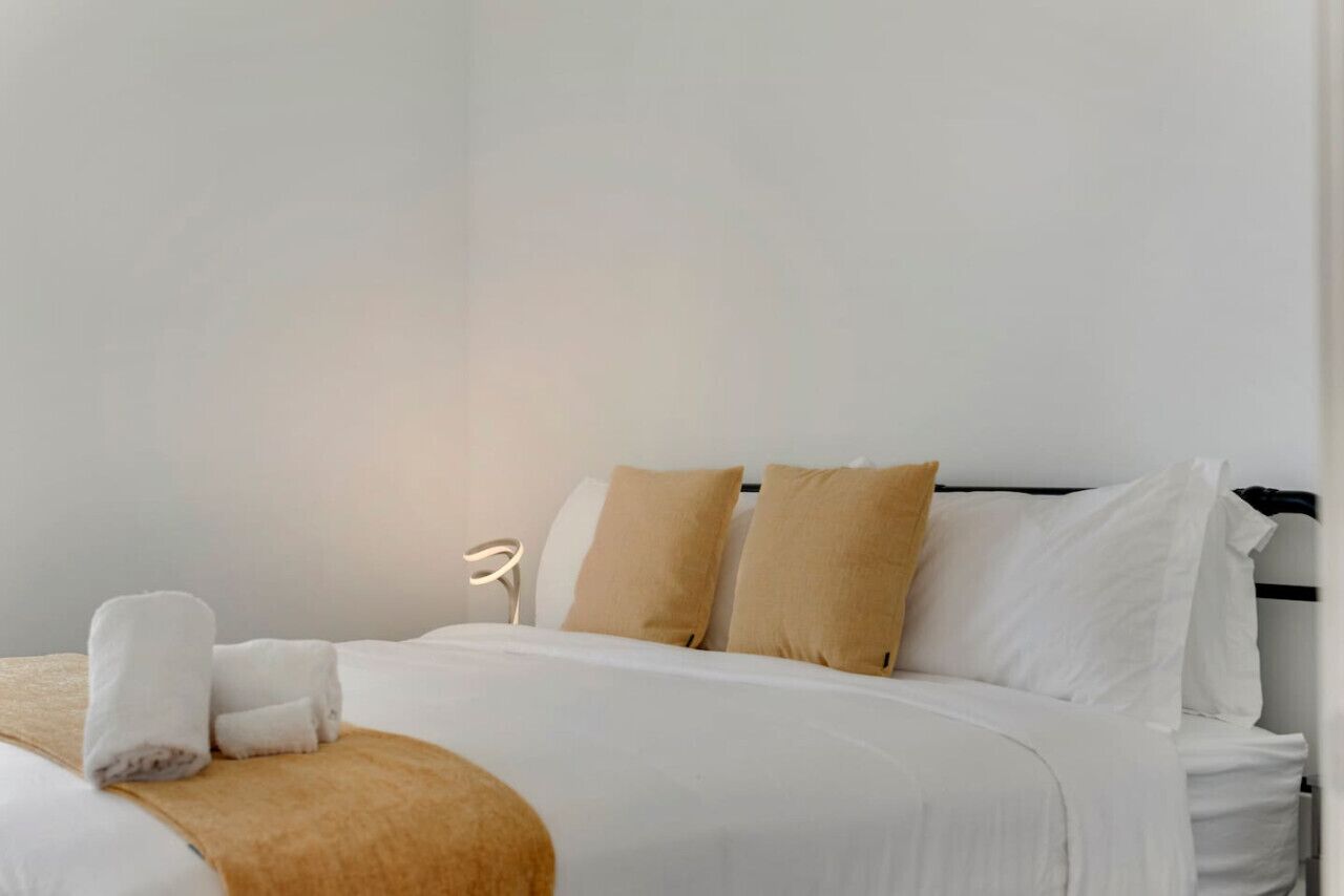 Un lit bien fait, avec une literie blanche et beige, une lampe de chevet et un luminaire fixé au mur.
