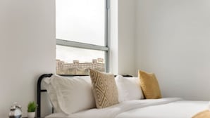 A bedroom with a bed, pillows, a nightstand, a plant, and a view of buildings through a window. - Modern 2BR | Gym | Parking | Workspace (Dallas)