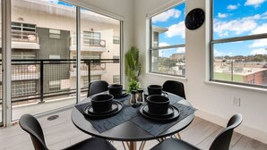 A dining area with a table set for four, large windows, and a view of buildings. - Modern 2BR | Gym | Parking | Workspace (Dallas)