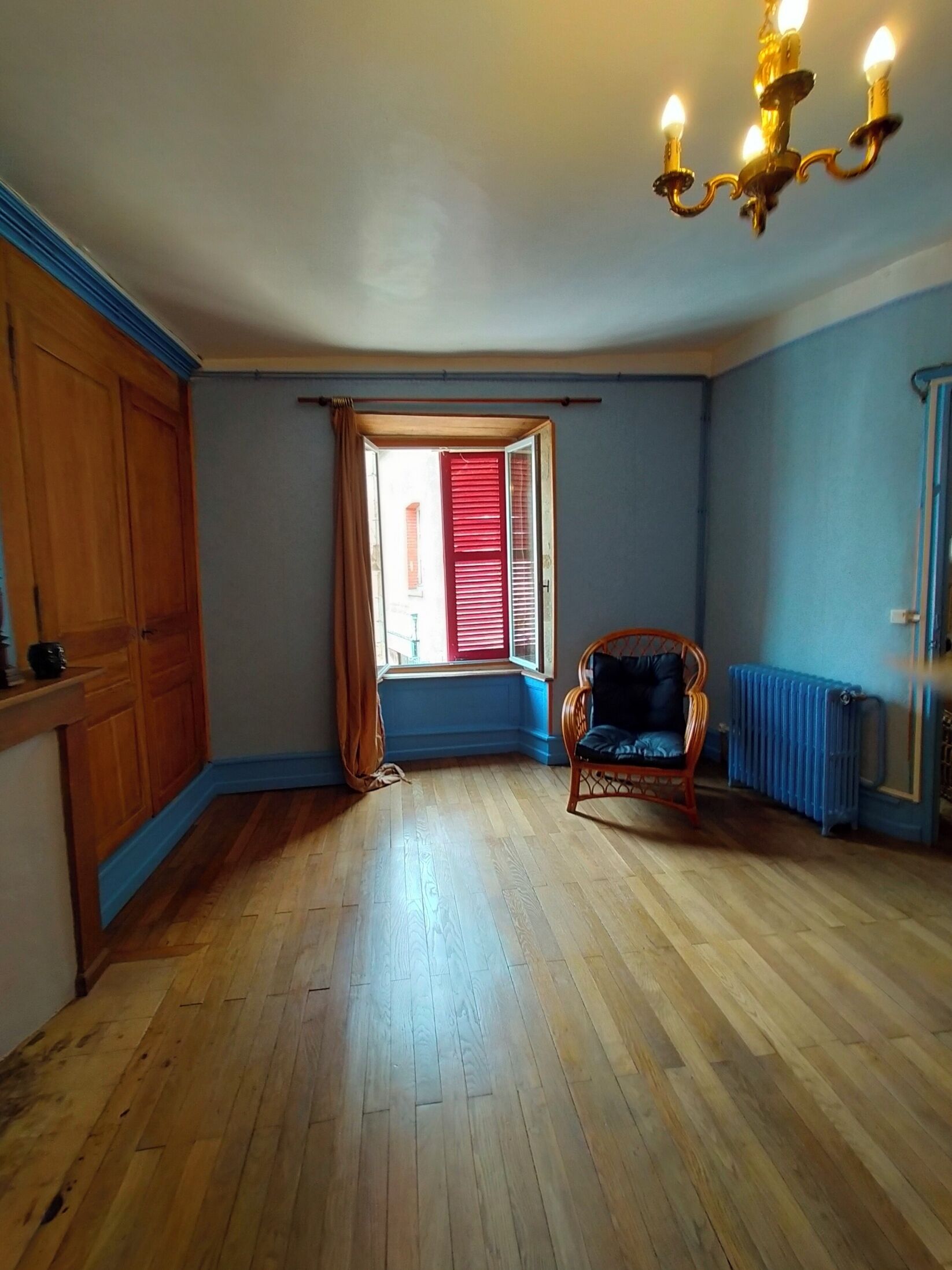 A room with wooden flooring, a window with red shutters, a wicker chair, and a radiator.