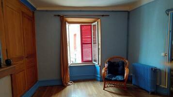 A room with wooden flooring, a window with red shutters, a wicker chair, and a radiator.