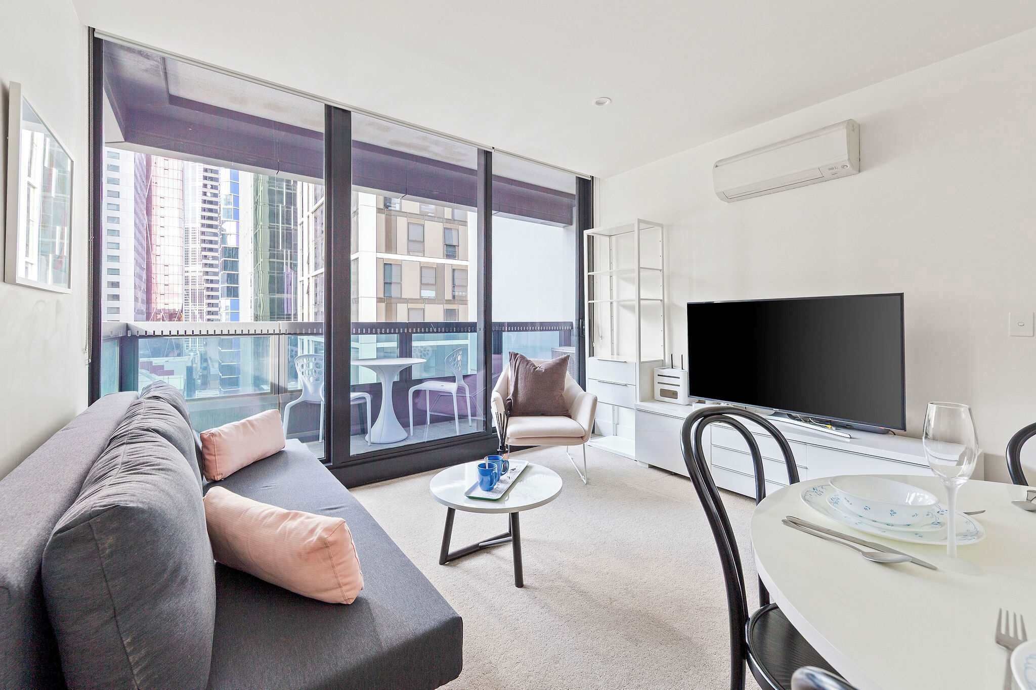 A modern living room with a sofa, a dining table set for two, a flat-screen TV, and a balcony with a view of high-rise buildings.