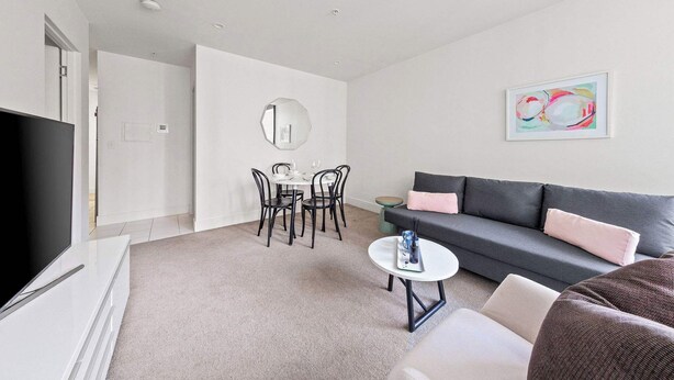 A modern living room with a flat-screen TV, a grey sofa, a round coffee table, and a dining area with a white table and black chairs. - City View 2BR, FREE Parking, Bright & Central Apt (Melbourne)
