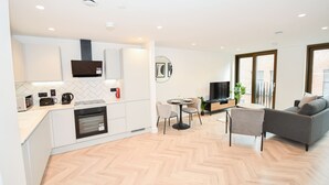 Cozinha moderna com armários brancos, forno embutido e micro-ondas. Há uma área de refeições com mesa redonda e cadeiras, e um sofá cinza na sala de estar.