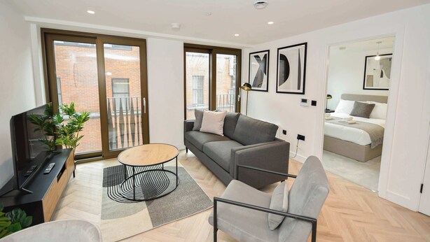 Uma sala de estar moderna com sofá, mesa de centro e aparador para TV. Possui uma varanda com vista para um edifício de tijolos.