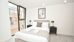 Quarto moderno com uma cama grande, duas mesas de cabeceira, uma obra de arte emoldurada e vista para um edifício de tijolos por uma janela ampla.