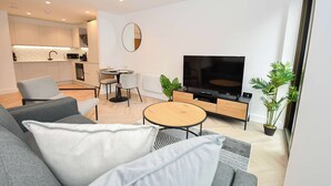 Uma sala de estar moderna com sofá, mesa de centro e televisão. Há uma área de cozinha com armários e mesa de jantar.