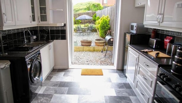 Une cuisine compacte avec des meubles blancs, un lave-linge et un coin repas visible à travers une porte ouverte.