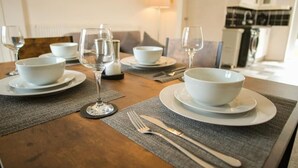 Une table à manger dressée avec des bols blancs, des verres à vin et des couverts, avec une cuisine en arrière-plan.