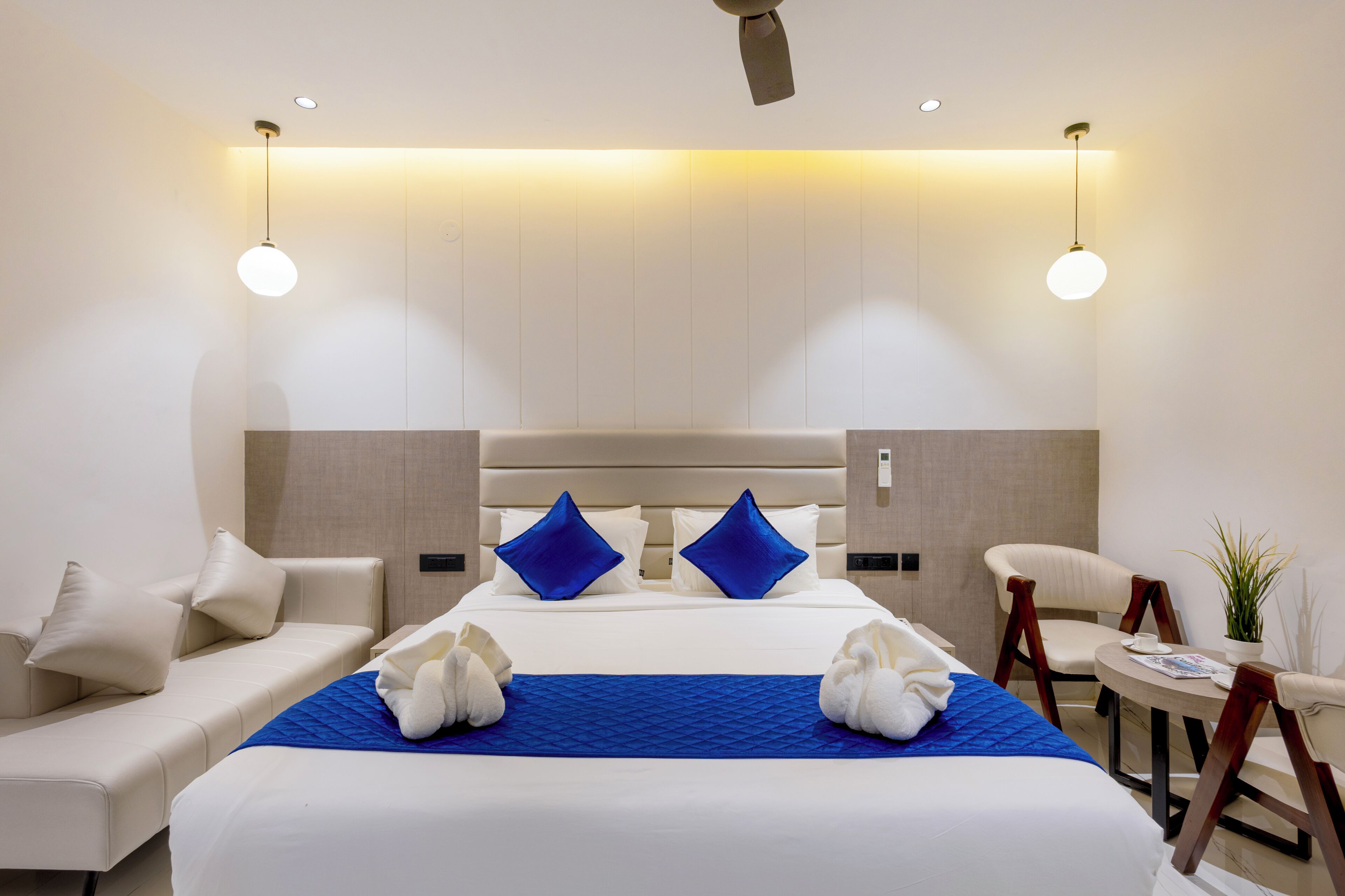 A hotel room with a large bed, two pillows, a blue bedspread, a white sofa, a chair, a small table, and a plant.