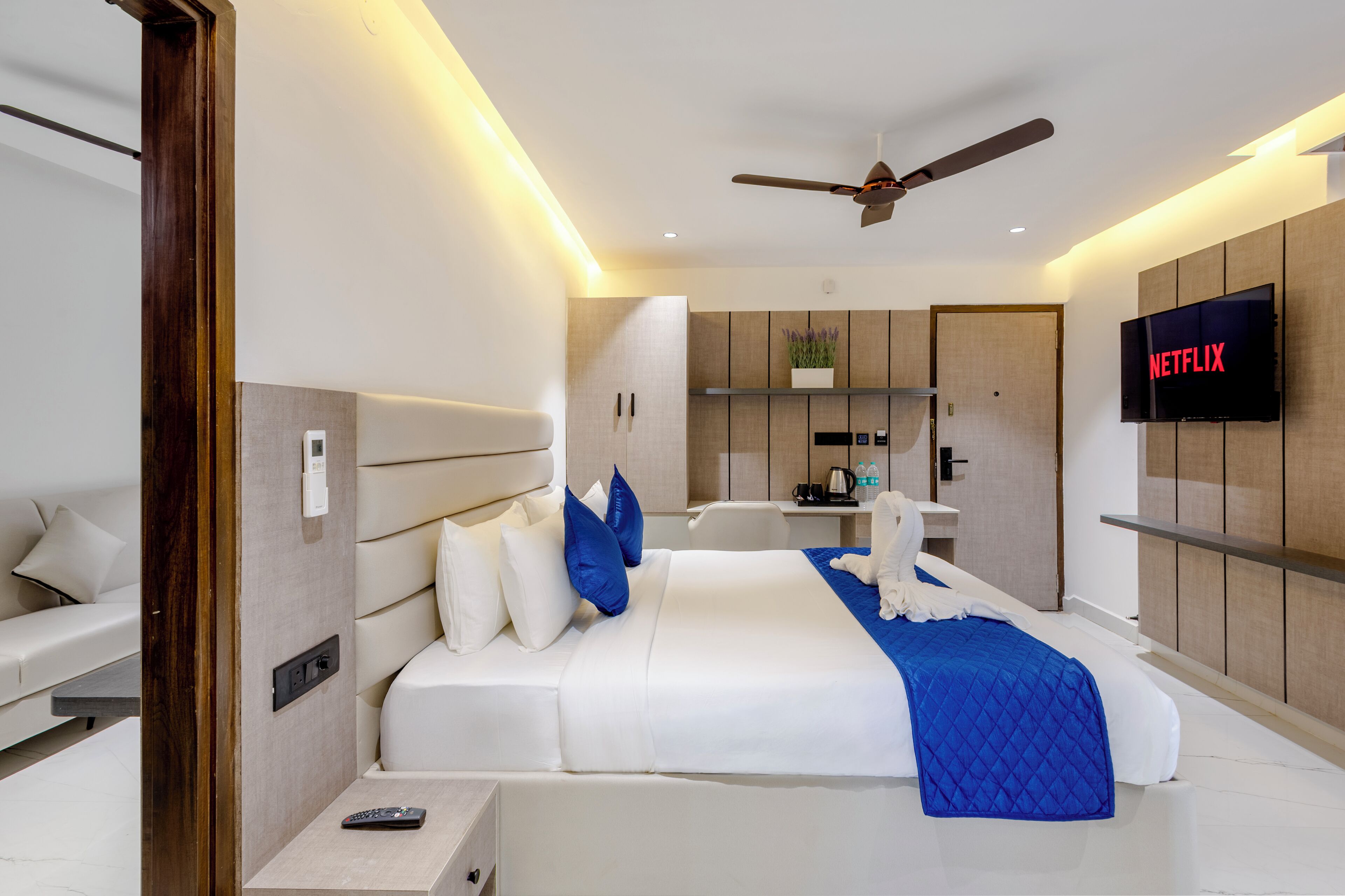 Habitación de hotel moderna con una cama grande, ventilador de techo, televisor montado en la pared y una pequeña kitchenette.
