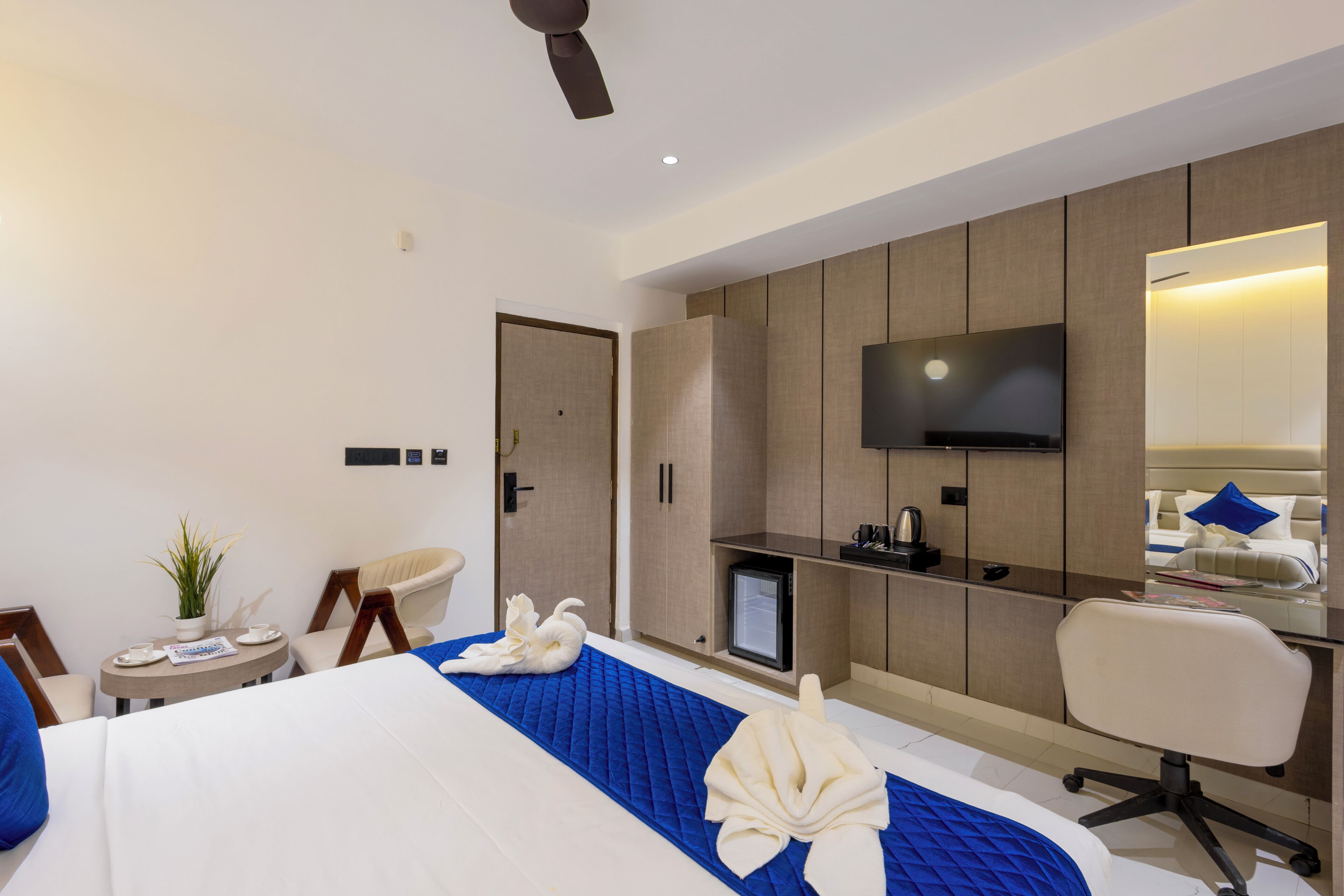 A modern hotel room with a bed, a desk, a chair, a TV, and a small table with a plant.