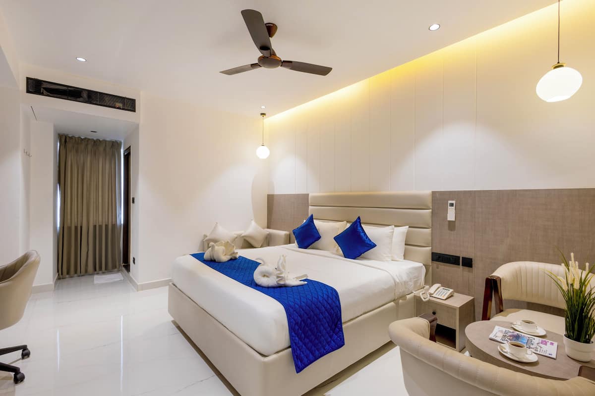 Une chambre d’hôtel moderne équipée d’un grand lit, d’un bureau, d’une chaise, d’un ventilateur de plafond et d’une petite table agrémentée d’une plante et d’un magazine.