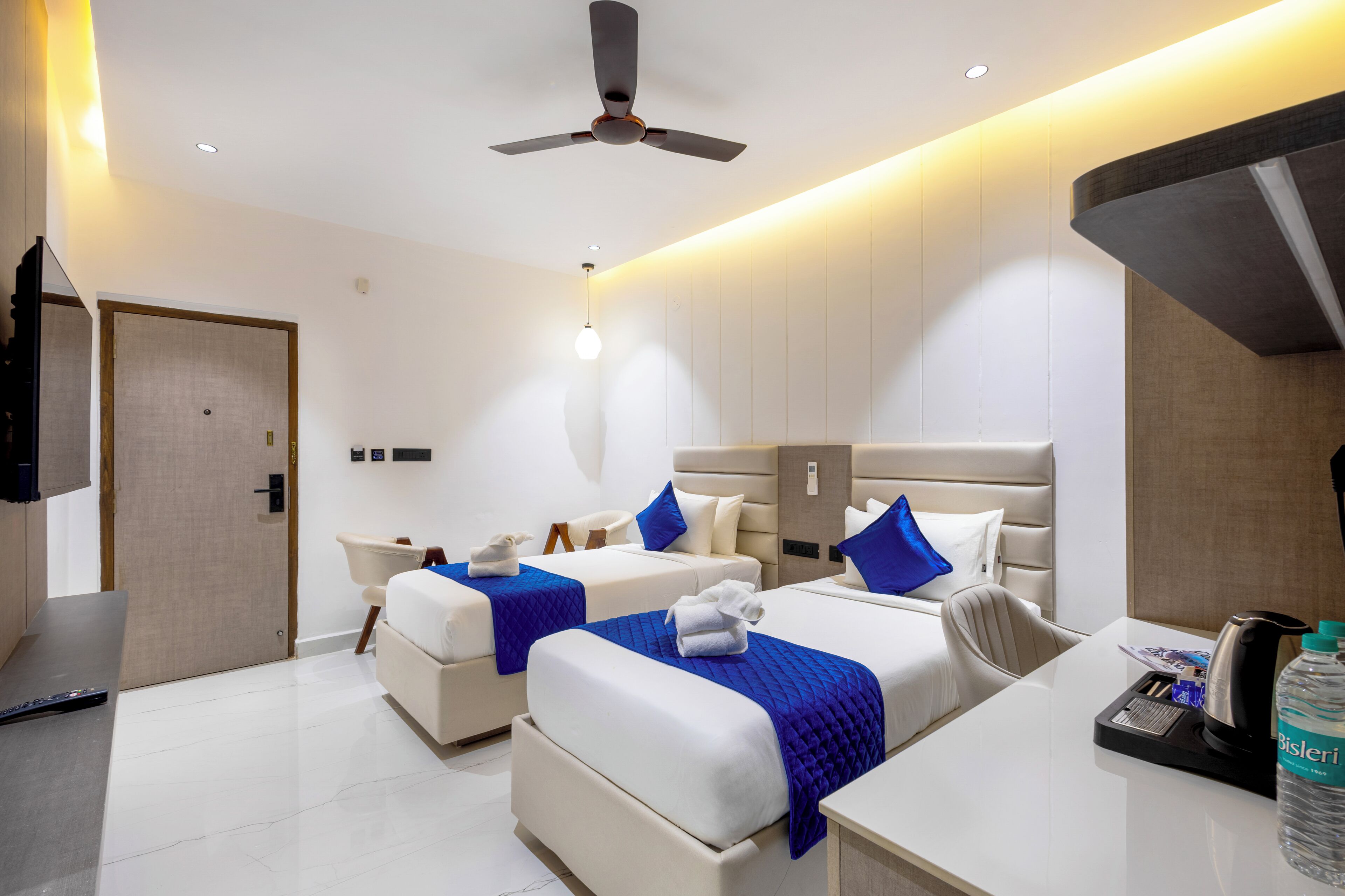 A modern hotel room with two beds, a desk, a chair, a ceiling fan, and a minibar.